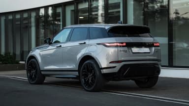 Rear three-quarter view of the silver crossover by a glass facade. Car 89