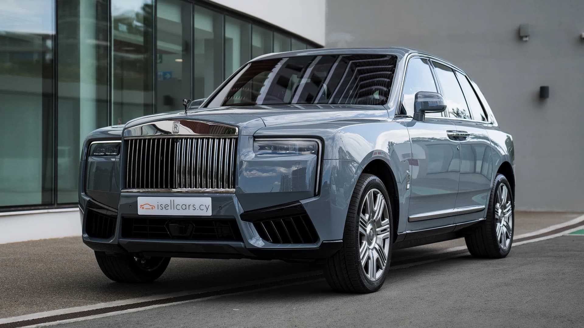 Front three-quarter view of a pale blue Rolls-Royce Cullinan outside a glass-fronted building